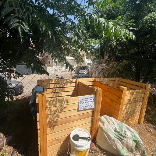 קומפוסטר עץ בגינה קהילתית לצד אדניות פורחות.